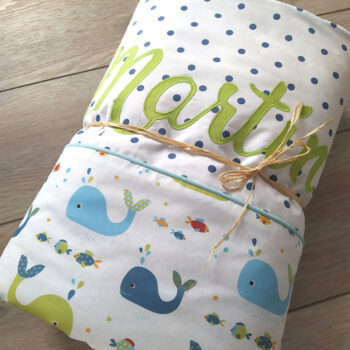 couverture pour bébé garçon personnalisé avec le prénom martin au tons vert et bleu, tissu blanc à pois bleu et tissu blanc à motif marrin, la couverture est présentée avec un lien en rafiat qui la tient roulée une création 2 Pommes et un mammouth