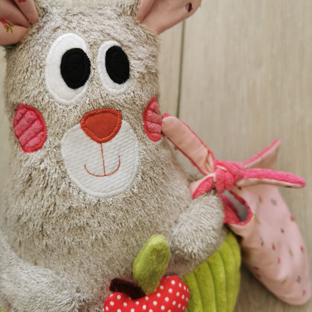 peluche musicale pour bébé fabriquée en France de manière artisanale, doudou dodu musical en éponge beige, aux joues rouges et au oreilles roses. il y a une petite pomme rouge à tirer sur son ventre pour lancer la musique. une création 2 pommes et un mammouth