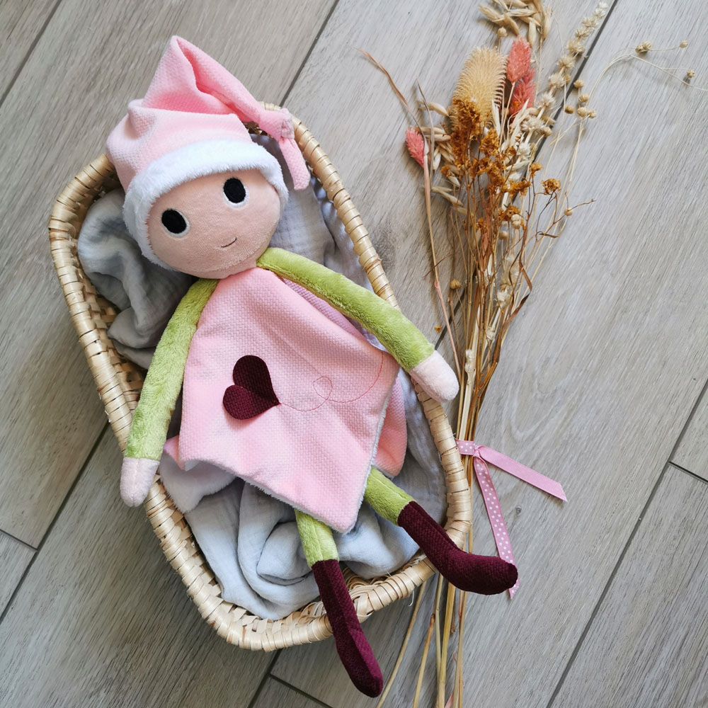 doudou pour bébé fabriqué en France de manière artisanale lutin patata présentée dans un corbeille en osier avec un bouquet de fleur séchées à droite. ce lutin a sa robe et son bonnet qui sont rose pâle, ses bras et son corps sont vert, ses bottes sont bordeaux. un cœur bordeaux est cousu sur sa robe, une création 2 pommes et un mammouth