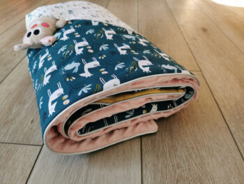 couverture pour petite fille présentée roulée , tissu minky rose poudrée, tissu coton bleu motif animaux de la forêt et tissu assorti blanc avec des oiseaux, une poche secret laisse dépasser un doudou dodu, une création 2 pommes et un mammouth