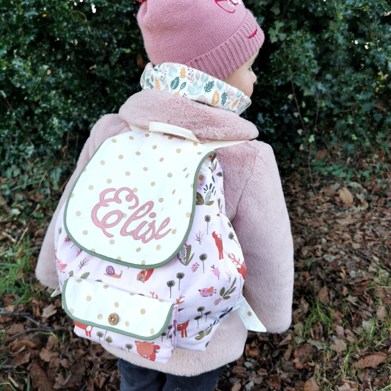 sac à dos maternelle personnalisé au prénom Elise, fabriqué en france de manière artisanale il est rose avec des animaux de la forêt et porté sur le dos d'une petite fille, une création 2 pommes et un mammouth