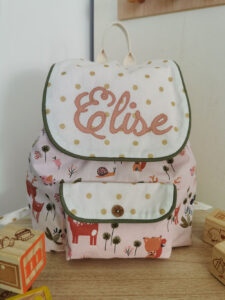 sac à dos maternelle personnalisé au prénom Elise, la poche principale et la poche de devant sont en tissu coton rose avec des animaux de la forêt, les rabats des poches sont blancs avec des pois dorés, tout autour est monté un passe poil vert kaki, sur la poche de devent et cousu un bouton en bois, une création 2 pommes et un mammouth