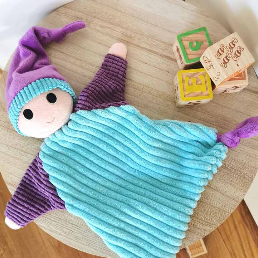 doudou pour bébé fabriqué en France de manière artisanale lutin patati posé sur une panière avec le couvercle en bois. la photo est prise du dessus, on voit le lutin en entier, son corps bleu turquoise en tissu grosses côtes, ses bras gaufrés violets et son bonnet violet assorti à ses pieds noués. Seule sa tête est rembourrée, il a de grand yeux ronds et un petit sourire, à ses côtés une pile de 4 cubes en bois, une création 2 pommes et un mammouth