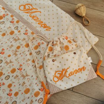 couverture et sac à dos ficelle personnalisé prénom manon, tissu coton blanc avec les animaux de la forêt en orange assorti à un coton blanc à pois dorés, le prénom est écrit en orange 2 pommes et un mammouth