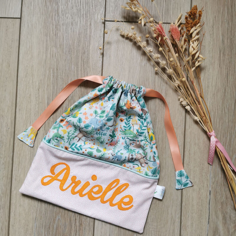 pochon personnalisé prénom Arielle, on retrouve un tissu bleu clair avec un motif jungle coloré et un tissu blanc à points roses sur lequel le prénom est en tissu orange cousu, le pochon se ferme par deux rubans en satin rose, une création 2 pommes et un mammouth