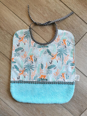 grand bavoir pour les repas de bébé présentée sur fond en bois , bavoir bi-matière en éponge de coton bleu turquoise et en tissu coton blanc motif forêt bleu et singes oranges, une petite dentelle vient réhaussée l'ensemble, une création 2 pommes et un mammouth