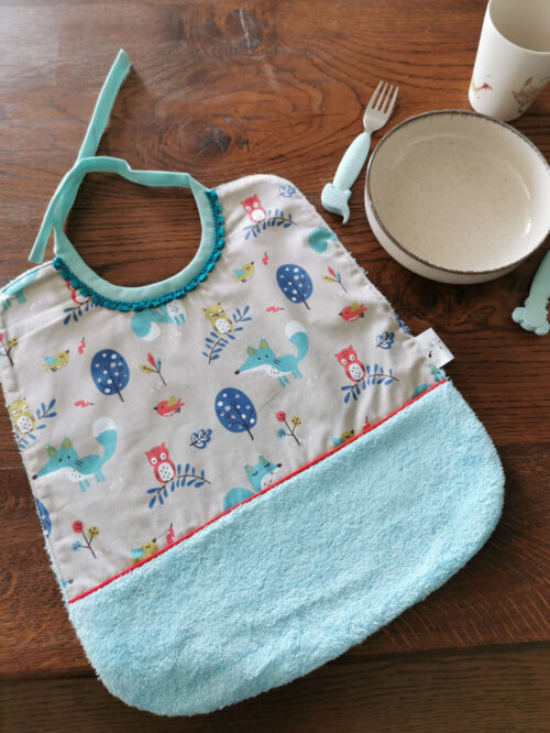 grand bavoir pour les repas de bébé présentée sur un table en bois avec une assiette et des couverts pour enfant sur un côté, bavoir bi-matière en éponge de coton bleu turquoise et en tissu coton gris avec des animaux de la forêt rouge et bleu , une petite dentelle vient réhaussée l'ensemble, une création 2 pommes et un mammouth