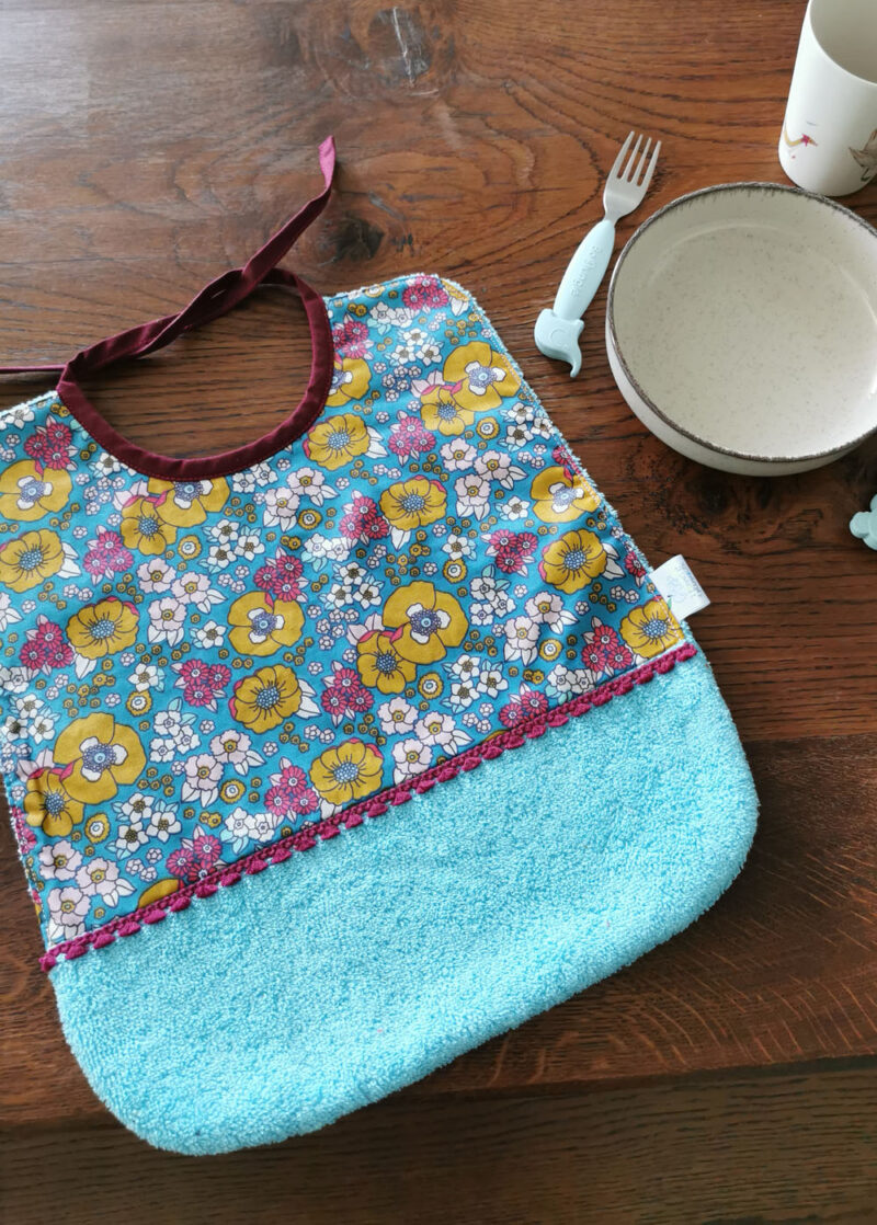 grand bavoir pour les repas de bébé présentée sur un table en bois avec une assiette et des couverts pour enfant sur un côté, bavoir bi-matière en éponge de coton bleu turquoise et en tissu enduit bleu fleuris , une petite dentelle vient réhaussée l'ensemble, une création 2 pommes et un mammouth