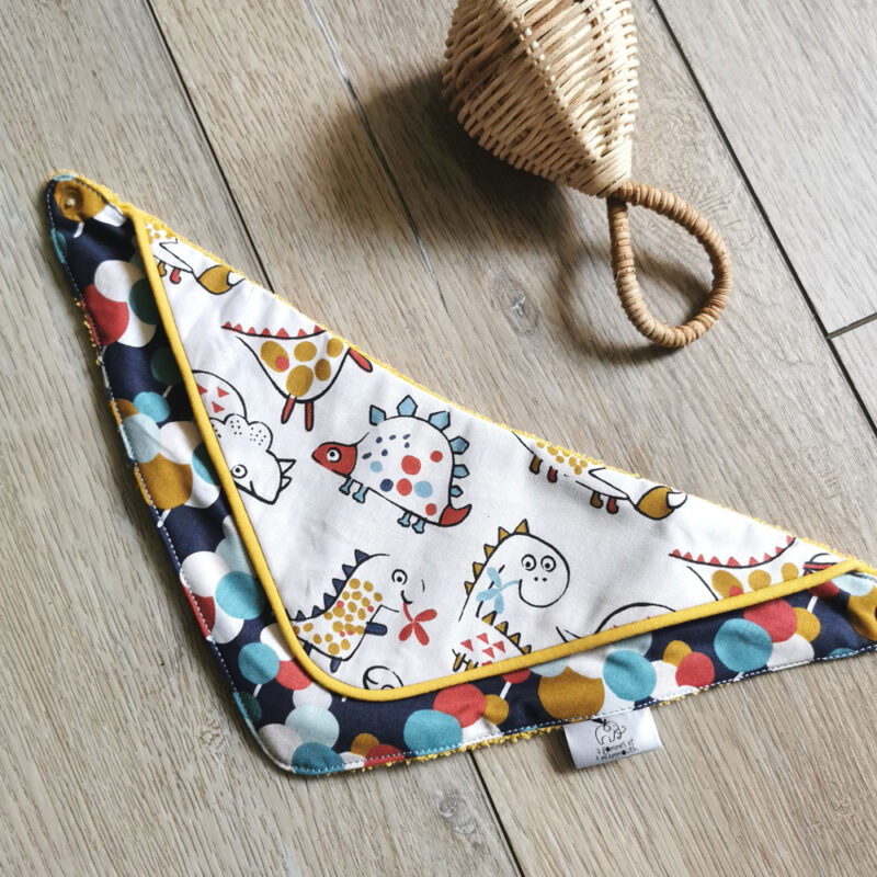 bavoir bandana anti-bave pour bébé, présenté avec un hochet en osier sur un fond bois bavoir bi-matière en éponge de coton jaune et en tissu coton blanc avec des dinosaures et un tissu coton bleu marine à pois rouges et bleus, un passepoil vient réhaussée l'ensemble, une création 2 pommes et un mammouth