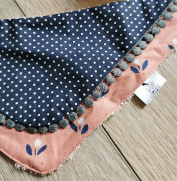 bavoir bandana anti-bave pour bébé, présenté avec un hochet en osier sur un fond bois bavoir bi-matière en éponge de coton jaune et en tissu coton bleu marine à petits pois blanc et un tissu coton rose brique à fleurs bleus, une dentelle grise vient réhaussée l'ensemble, une création 2 pommes et un mammouth