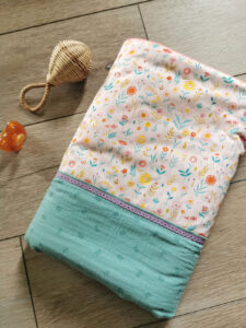 couverture pour bébé fille présentée roulée aux couleurs estivales, tissu vert d'eau et tissu fleuri sont réhaussés d'une dentelle parme et d'un liseret orange, une création 2 pommes et un mammouth