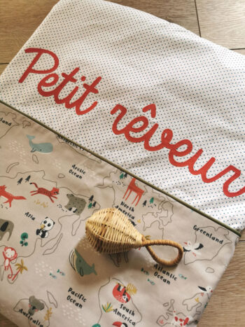 couverture enfant personnalisée Petit rêveur aux couleurs tendre bleu kaki, taupe et brique, présentée pliée avec un hochet en osier plier dessus , une crétion 2 pommes et un mammouth