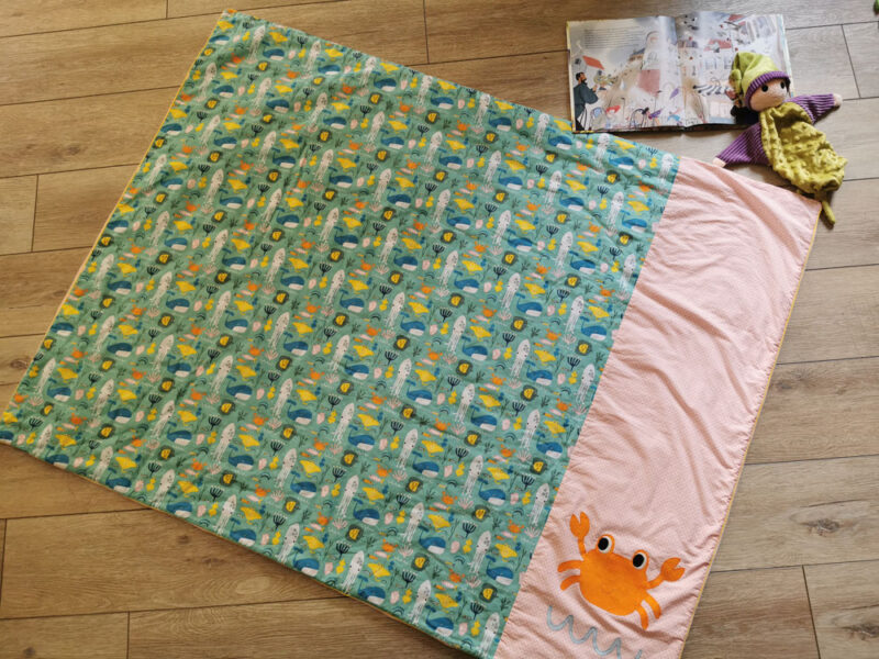 couverture pour enfant aux couleurs vives : rose, orange, bleu, jaune, tissus au motifs de la mer avec poissons, pieuvres et baleine, un gros crabe orange en appliqué, couverture présentée dépliée sur le sol , une création 2 pommes et un mammouth