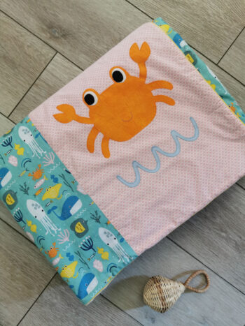 couverture pour enfant aux couleurs vives : rose, orange, bleu, jaune, tissus au motifs de la mer avec poissons, pieuvres et baleine, un gros crabe orange en appliqué, couverture présentée pliée, une création 2 pommes et un mammouths