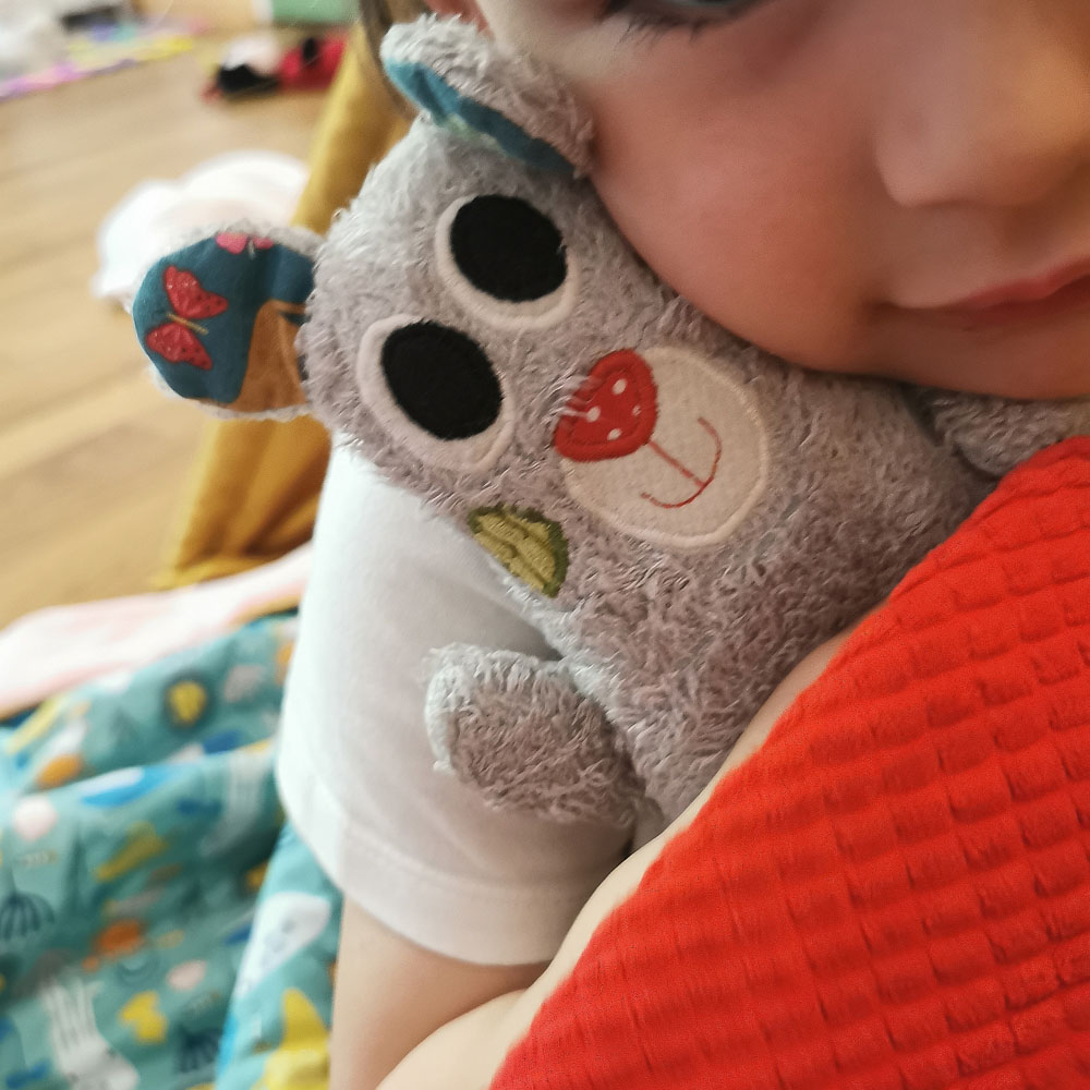 doudou pour bébé fabriqué en france de manière artisanale, un dodu qui se fait câliner par une petite fille, ce doudou est gris clair avec les oreilles verte et le nez rouge, il a de grands yeux expressifs et un petit sourire. une création 2 pommes et un mammouth