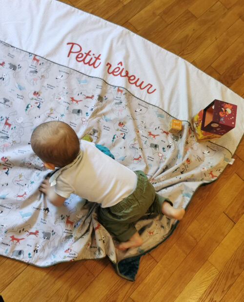 couverture géante pour enfant fabriqué en france de manière artisanale, elle est posée au sol et un bébé marche à quatre pattes dessus. Elle est beige avec des motifs de planisfères et les animaux du monde, elle est personnalisée au nom 