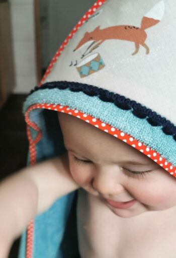 bébé qui sorrt du bain et qui a sur la tête la capuche de sa cape de bain 2 pommes et un mammouth, elle est en microéponge bleu, sur la capuche on trouve un tissu en coton beige avec un renard et un galon de pompons bleu marine. les finitions sont approtées par un biais orange avec des pois blancs