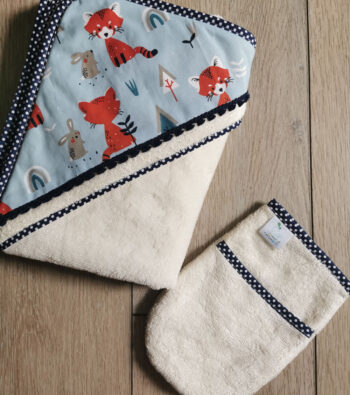 photo de la cape de bain pour bébé et de son gant malin assorti 2pommes et un mammouth, en microéponge blanche, un biais bleu marine à pois blanc est posé tout le tour, la capuche est en tissu coton bleu clair avec des renards orange et des lapins gris, elle est doublée en microéponge, un galon de pompons bleu marine est mis en décoration le long de la capuche. Le gant a une poche pour mettre le savon, il est en microéponge blanche rehausse d'un biais bleu marine à pois blancs. L'ensemble est présenté posé sur un fond en bois.