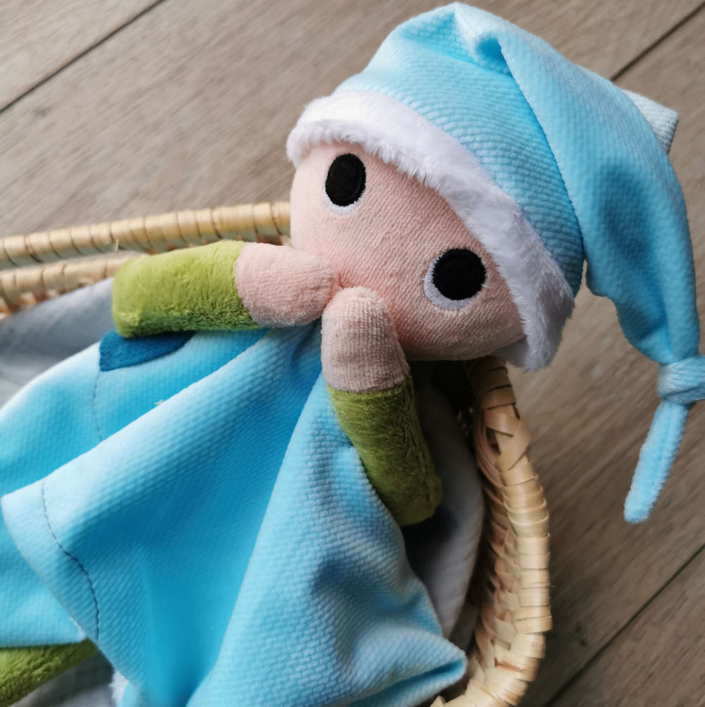 doudou pour bébé fabriqué en France de manière artisanale lutin patata , il est installée dans une panière en osier, les deux mains sur la bouche, il a un regard très expressif avec ses yeux ronds. Sur sa tête un bonnet bleu clair, ses bras sont verts et il a une "robe" en forme d'étoile assorti à son bonnet. Un petit cœur bleu foncé est cousu sur sa robe. une création 2 pommes et un mammouth